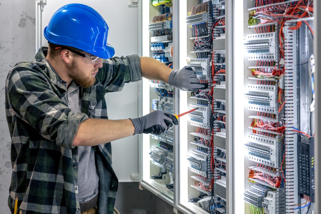 Electrician wearing helmet and gloves installs cables in an electrical switchboard using a screwdriver and technical tools.